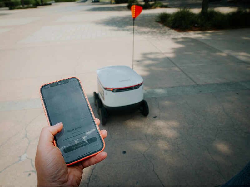 Person with a smartphone behind a last-mile delivery robot traveling down a street